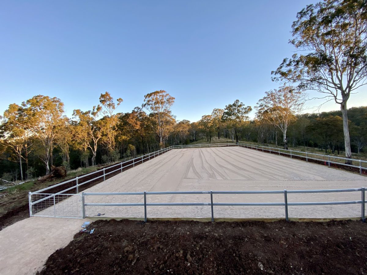 A large, fenced sand arena surrounded by trees under a clear sky at dusk.